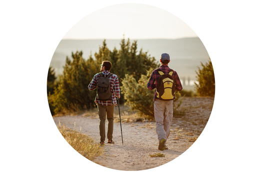 Two people hiking on a dusty trail through a dry landscape, walking away from the camera with backpacks and trekking poles at golden hour.